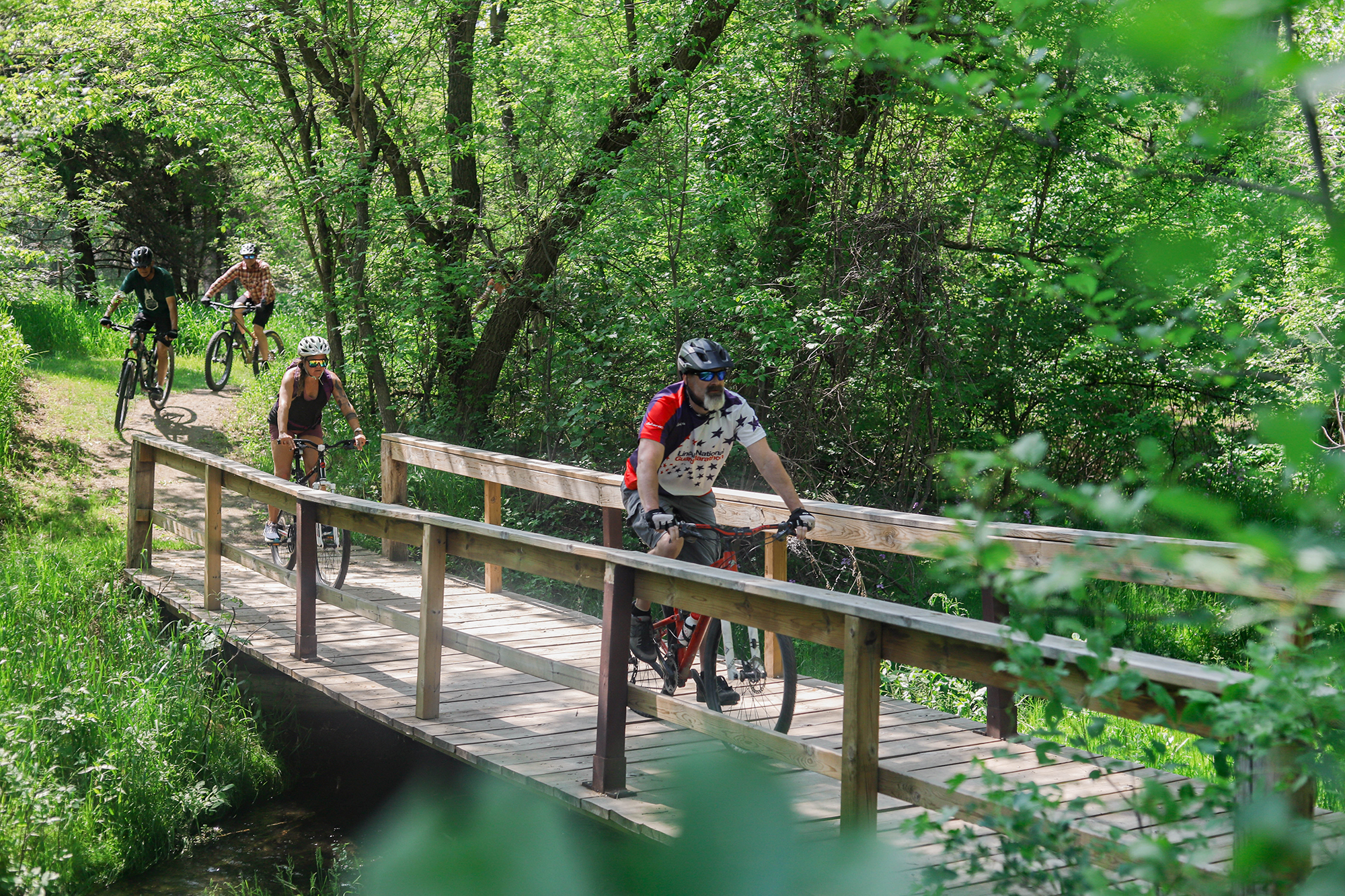 Cyclists crossing a bridge surrounded by green vegetation in Chadron State Park.