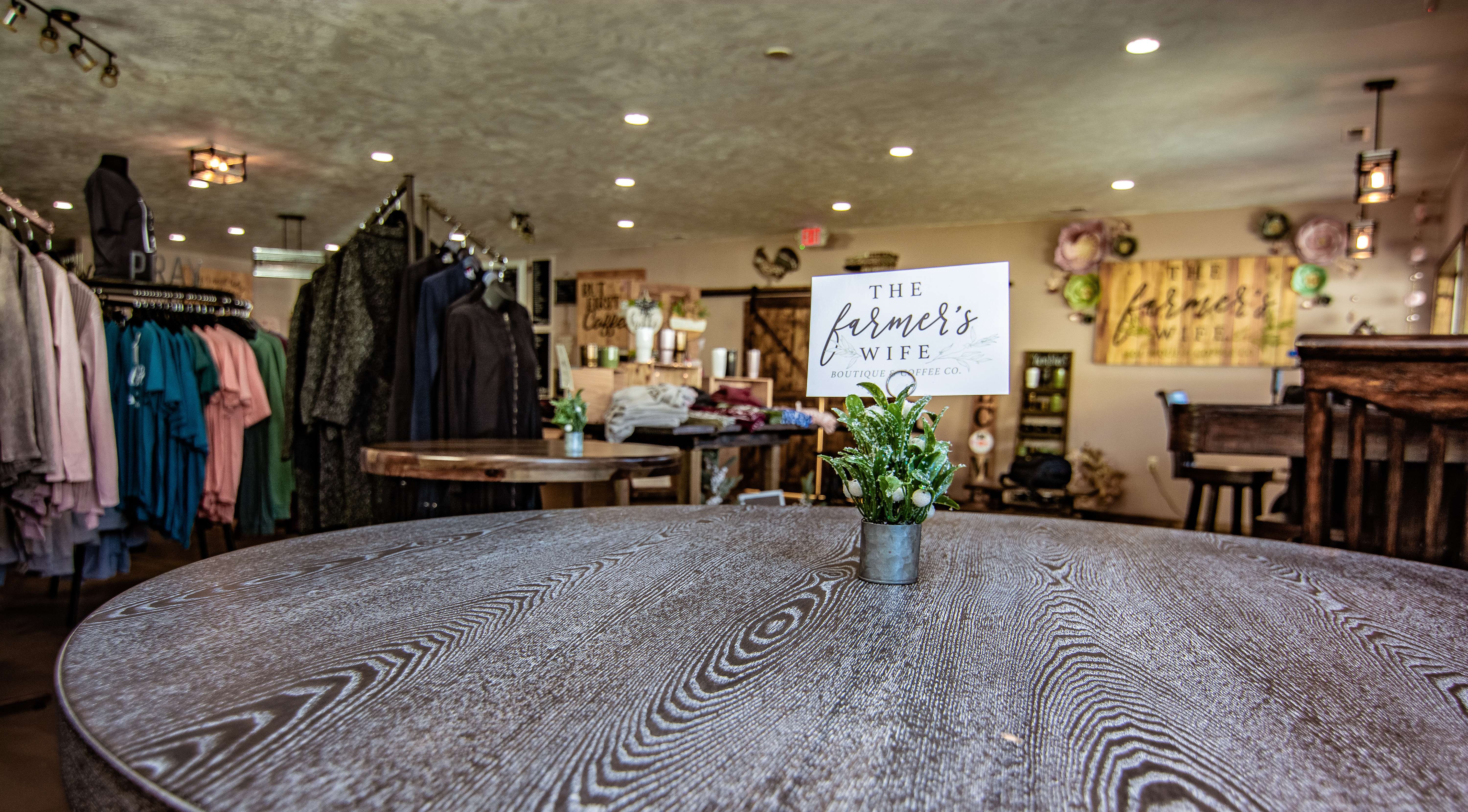 Clothing racks and a sign reading "The Farmer's Wife Boutique & Coffee Co." on a centerpiece inside the boutique..